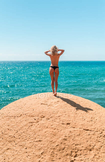 Полный вид назад на неузнаваемую топлесс-блондинку в брюках, стоящую на каменистом побережье против спокойного бирюзового моря и наслаждающуюся солнечным днем во время летних каникул — стоковое фото