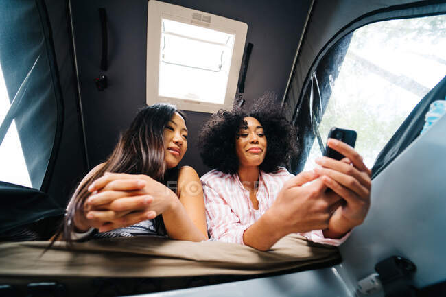 De baixo de positivo jovem asiático e afro-americano amigos do sexo feminino navegando telefone celular enquanto descansando juntos dentro van campista durante a viagem de verão na natureza — Fotografia de Stock