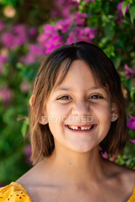 Fröhliches Kind mit braunen Augen in gelben Klamotten blickt tagsüber in die Kamera gegen blühenden Strauch — Stockfoto