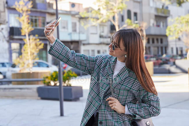 Jovem elegante fêmea na moda houndstooth casaco e óculos de sol tomando selfie no smartphone, enquanto de pé na rua da cidade — Fotografia de Stock