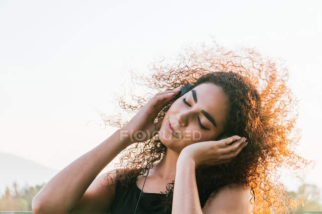 Unbekümmerte junge hispanische lockige Frau hört Musik über Kopfhörer, während sie an sonnigen Sommerabenden auf der Straße chillt — Stockfoto