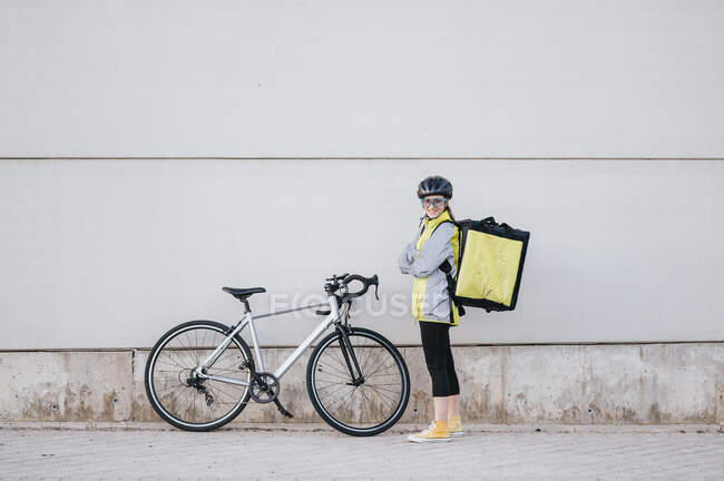 Full body confident female courier with thermal bag crossing arms and looking at camera with smile while standing near bike outside modern building on city street — Stock Photo