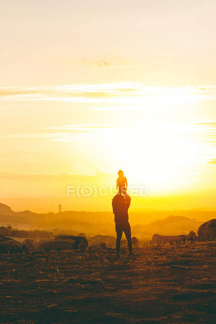 Visão traseira do homem anônimo que levanta a criança no ar no vale áspero rochoso no por do sol — Fotografia de Stock