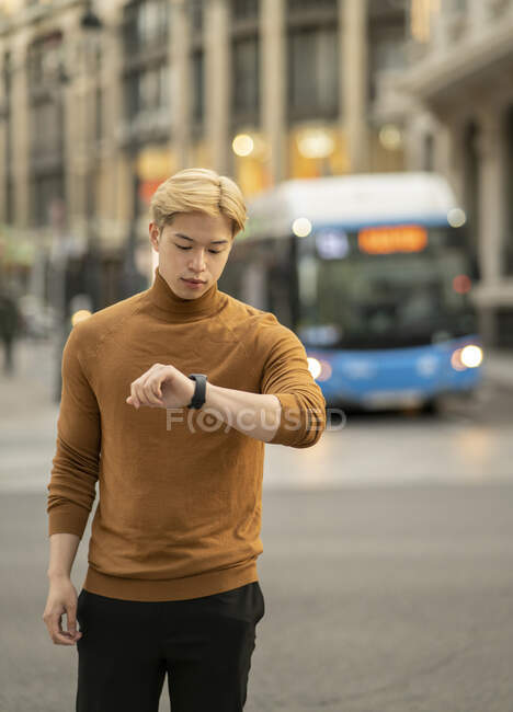 Serious ethnic male checking time on wristwatch while standing in street and waiting for meeting — Stock Photo