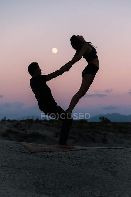Вид збоку силует невпізнаваної гнучкої жінки, що стоїть на ногах чоловіка під час сеансу акро-йоги на тлі вечірнього неба — стокове фото