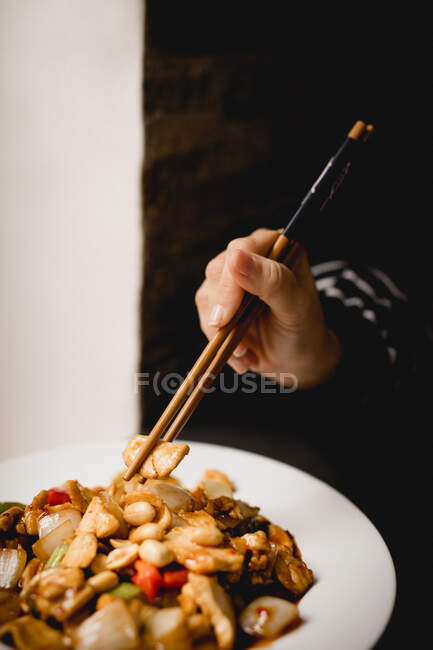 Mulher irreconhecível usando pauzinhos para comer porção de frango Gong Bao delicioso contra fundo preto no restaurante — Fotografia de Stock