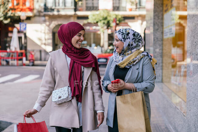 Amies musulmanes gaies avec des sacs en papier marchant en ville après avoir fait du shopping en se regardant — Photo de stock