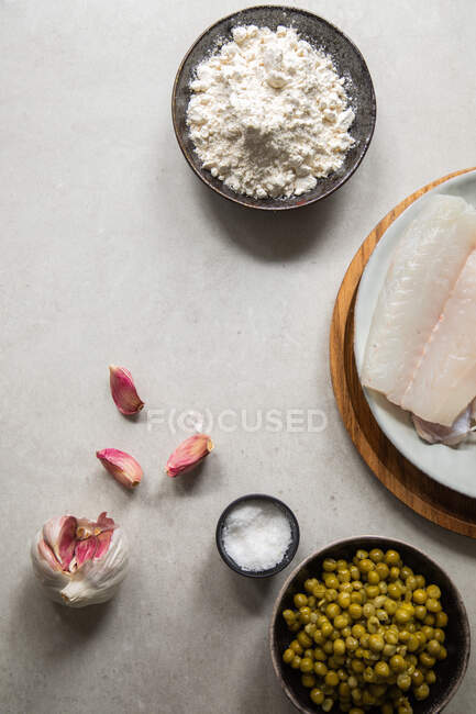 From above top view fresh garlic cloves and salt placed on table near hake fillet and bowl with peas during food preparation in kitchen — Stock Photo