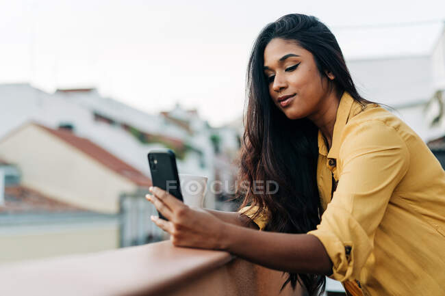 Giovane donna ispanica gustare caffè fresco e utilizzando il telefono cellulare mentre si rilassa sul balcone in serata — Foto stock