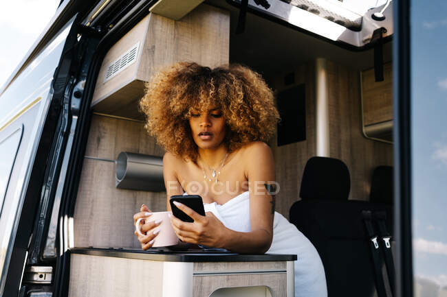 Mulher afro-americana feliz com caneca de bebida quente sorrindo e navegando celular enquanto descansa na caravana contemporânea pela manhã — Fotografia de Stock