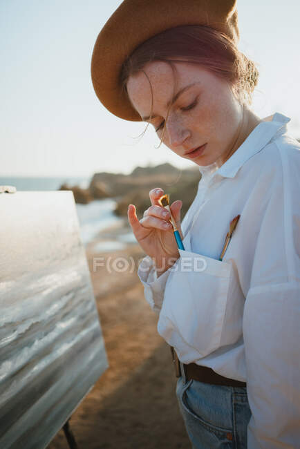 Side view of young thoughtful female artist in stylish outfit and beret standing on shore near sand and ocean while drawing seascape on canvas on easel in sunny day with paintbrush — Stock Photo