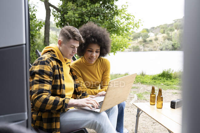 Young diverse man and woman in casual clothes smiling and browsing netbook while sitting near van during road trip in nature — Stock Photo