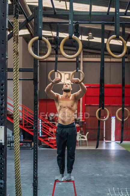 Longitud completa hombre sin camisa fuerte de pie en el taburete y preparándose para hacer ejercicio en anillos gimnásticos durante el entrenamiento intenso en el gimnasio moderno - foto de stock