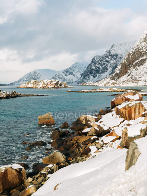 Rocas nevadas y crestas de montaña ubicadas en la costa cerca del mar ondulante contra el cielo nublado en invierno en las Islas Lofoten, Noruega - foto de stock