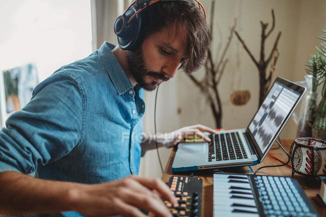 Вид збоку молодого чоловіка в навушниках, використовуючи синтезатор і ноутбук за столом вдома — стокове фото
