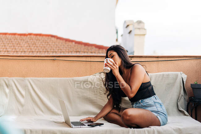 Joven mujer hispana bebiendo café caliente de la taza y navegando en netbook trabajando remotamente mientras está sentada con las piernas cruzadas en el sofá por la mañana en el balcón - foto de stock