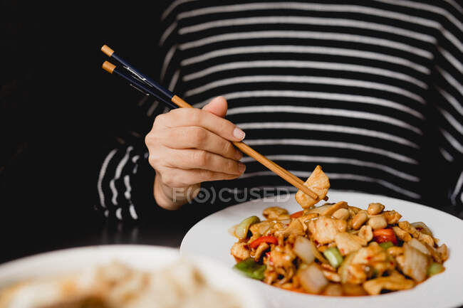 Unerkennbares Weibchen mit Essstäbchen isst Portion leckeres Gong Bao Huhn vor schwarzem Hintergrund im Restaurant — Stockfoto