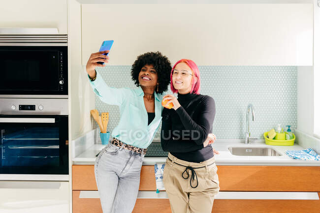 Joyful elegantes amigas multirraciales en traje casual de pie en la cocina casera y tomar selfie con teléfono móvil - foto de stock