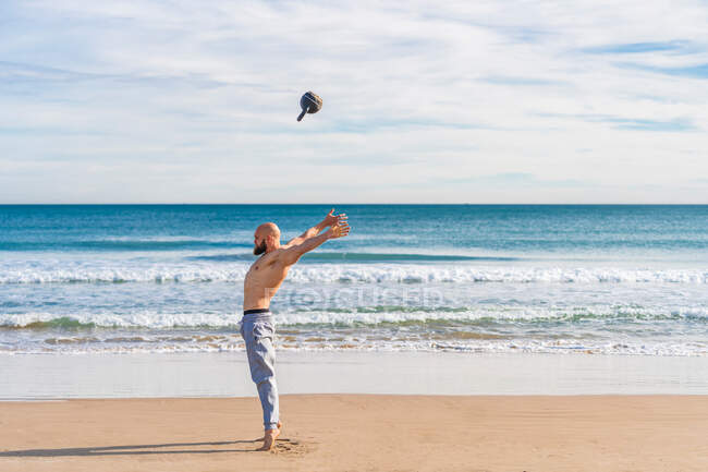 Vista lateral del hombre deportivo sin camisa lanzando kettlebell en la costa arenosa con olas del océano en el fondo - foto de stock