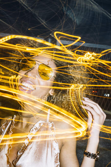 Mujer afroamericana joven y segura de sí misma con cabello rizado en gafas de sol de moda y top relajante en la calle por la noche cerca de luces de congelación - foto de stock