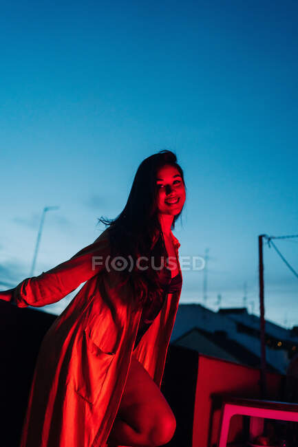 Encantada joven hembra en lencería sonriendo y mirando a la cámara mientras descansa en la terraza bajo la luz de neón roja por la noche - foto de stock