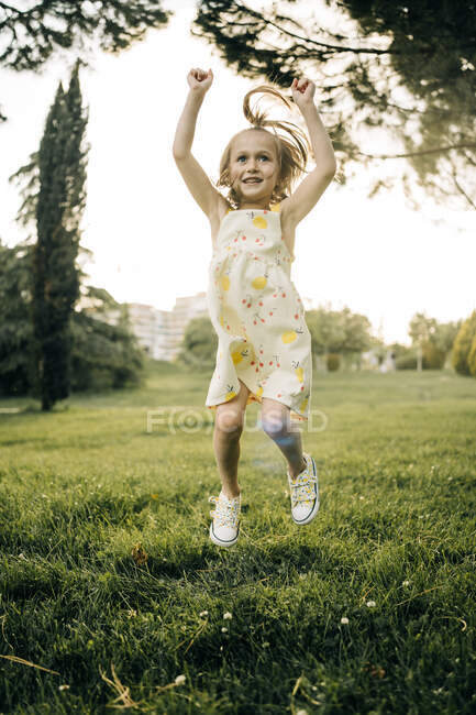Corps complet de joyeuse petite fille énergique en robe de soleil sautant haut au-dessus du sol tout en s'amusant dans un parc verdoyant en journée d'été — Photo de stock