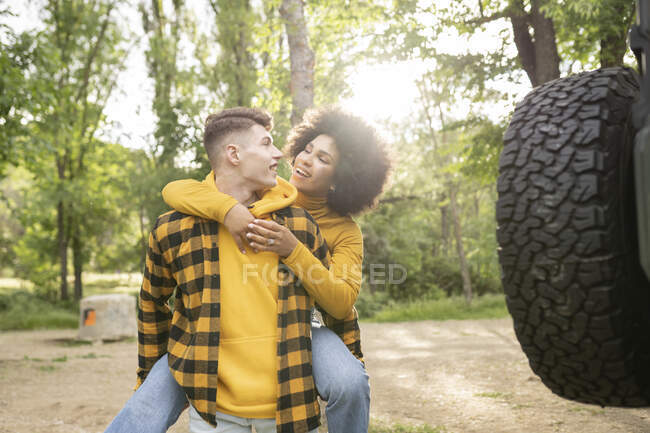 Молодой человек катается на спине к оптимистичной черной подруге повседневной одежде, стоя возле машины в солнечный день в сельской местности — стоковое фото