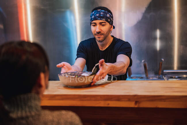 Homem barbudo em bandana e camiseta preta colocando placa com macarrão no balcão de madeira para o cliente em barra de ramen — Fotografia de Stock