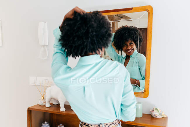 На задньому плані - вродлива молода афроамериканка в повсякденному одязі, що торкається кучерявого волосся, стоячи в кімнаті і дивлячись на дзеркало. — стокове фото