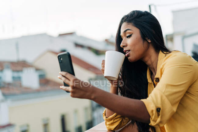 Giovane donna ispanica gustare caffè fresco e utilizzando il telefono cellulare mentre si rilassa sul balcone in serata — Foto stock