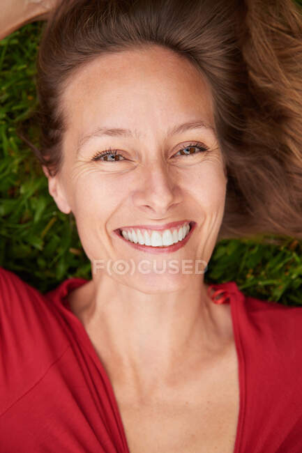 Mujer vestida de rojo tendida en el suelo en un parque con hierba y mirando a la cámara - foto de stock
