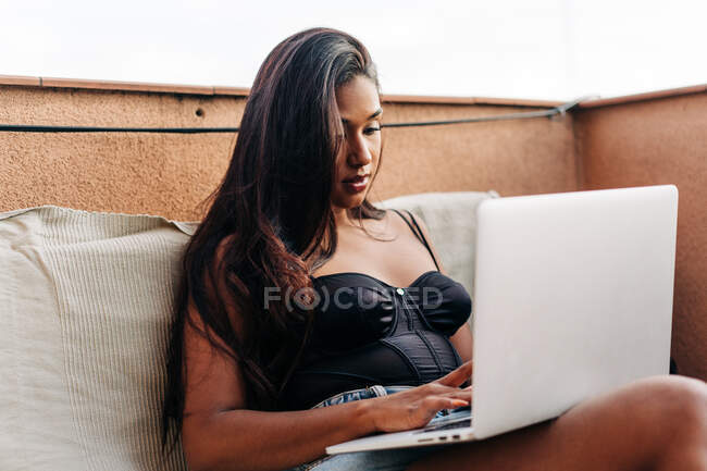 Молода іспаномовна жінка з довгим волоссям сидить на дивані і переглядає соціальні мережі на ноутбуку, відпочиваючи на балконі у вихідні. — стокове фото