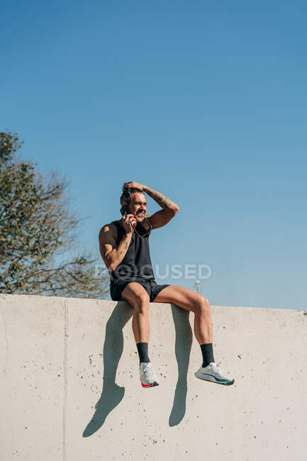 Fit male athlete in sports clothes talking on cellphone while resting on fence after workout — Stock Photo
