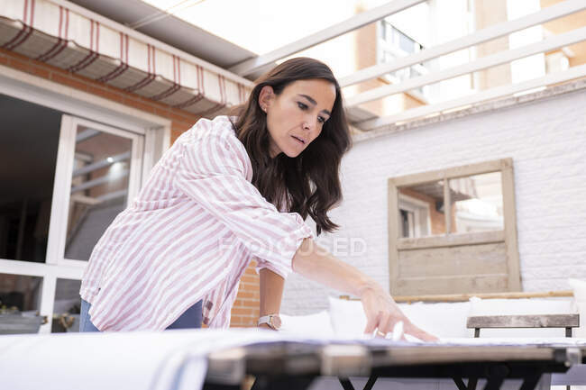 Positive female architect standing at table while analyzing and pointing at blueprint during work on project — Stock Photo