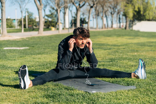 Atleta barbudo em sportswear e fones de ouvido sentado no prado na cidade — Fotografia de Stock