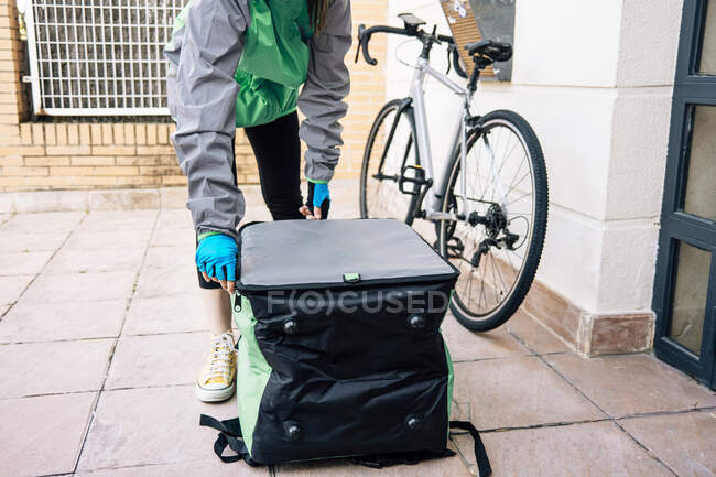 Anonymous female courier in uniform opening insulated thermal bag on tiled floor near door while making delivery — Stock Photo