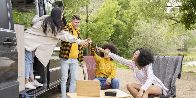 Group of multiracial travelers with beer gathering with beer near van and watching video on netbook during road trip in countryside — Stock Photo
