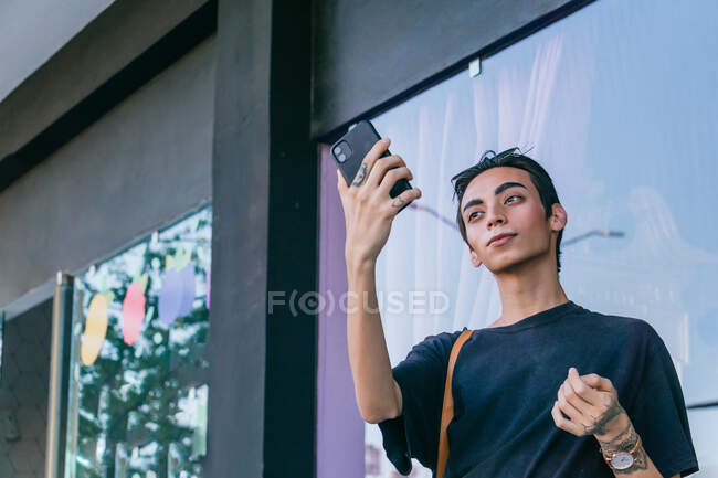 Низкий угол стильного гея, стоящего возле здания на улице и стреляющего в себя на смартфоне — стоковое фото