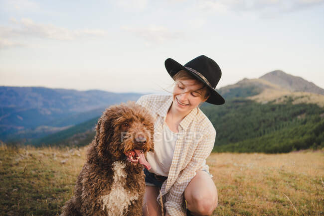 Alegre propietaria femenina con obediente perro Labradoodle sentado en las montañas mirando hacia otro lado - foto de stock