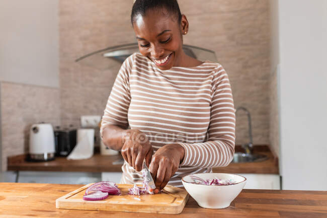 Врожай весела етнічна жінка, що ріже свіжу червону цибулю з ножем на обробній дошці за столом з мискою в будинку — стокове фото