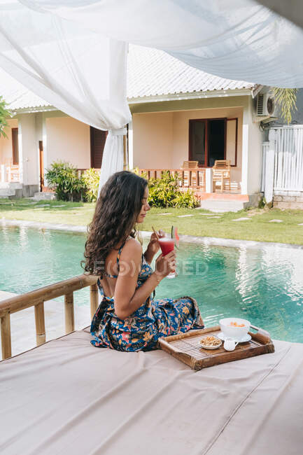 Back view of female tourist in sundress with floral ornament sitting with glass of refreshing drink on bed outdoors — Stock Photo
