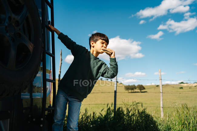 Cute little traveler biting tasty sandwich and looking away while hanging on ladder of modern RV on summer day in countryside — Stock Photo