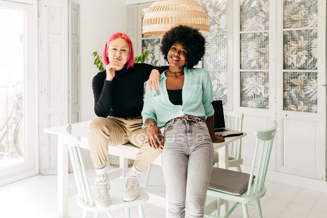 Joyeux jeunes amis féminins multiraciaux élégants regardant la caméra tout en étant assis ensemble sur la table dans la salle de lumière — Photo de stock