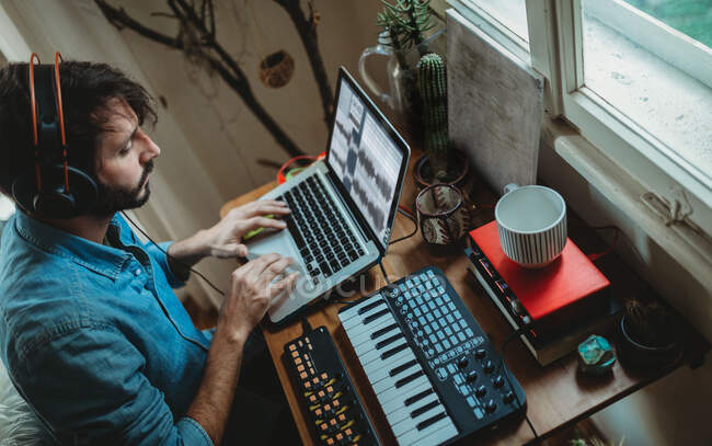 З іншого боку, сфокусований молодий чоловік у навушниках працює над синтезатором і ноутбуком удома. — стокове фото