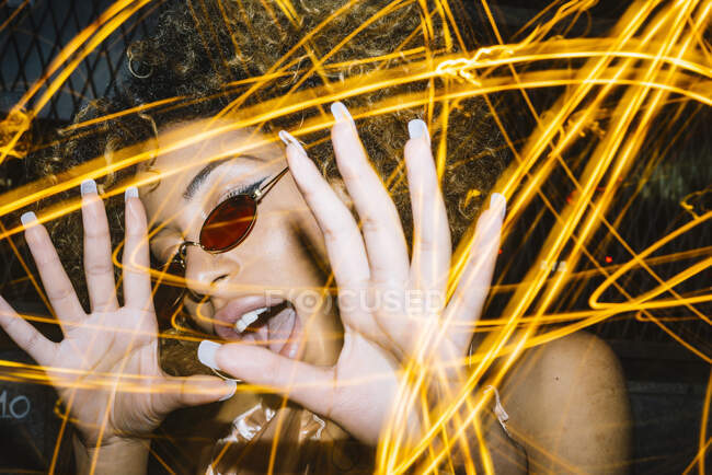 Jeune femme ethnique expressive avec des cheveux afro dans des lunettes de soleil élégantes et le dessus touchant la tête et criant bruyamment tout en refroidissant dans une boîte de nuit près des lumières de gel — Photo de stock