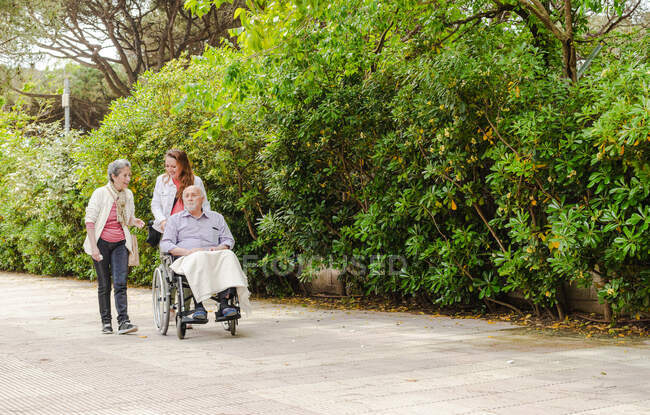 Senior mother and adult daughter walking with aged father in wheelchair in green summer park — Stock Photo