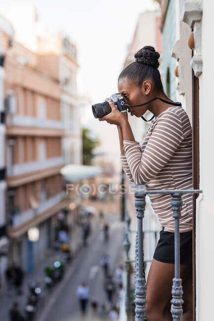 Вид сбоку этническая женщина в одежде с полосатым орнаментом с профессиональным фотоустройством глядя на балкон в дневное время — стоковое фото