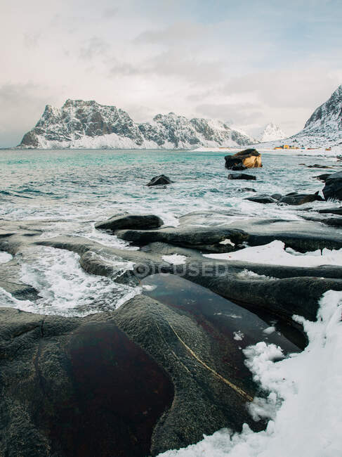 Água do mar fria salpicando em rochas perto da costa gelada e nevada perto de montanhas no dia de inverno cinza nas ilhas Lofoten, Noruega — Fotografia de Stock