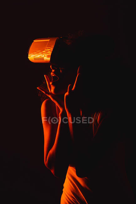 Mujer étnica anónima asombrada con boca abierta explorando realidad virtual en auriculares sobre fondo negro - foto de stock
