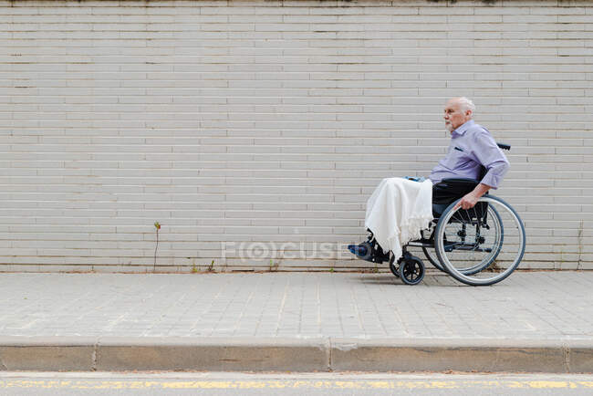 Side view of gray haired male in wheelchair riding along pavement in city — стоковое фото